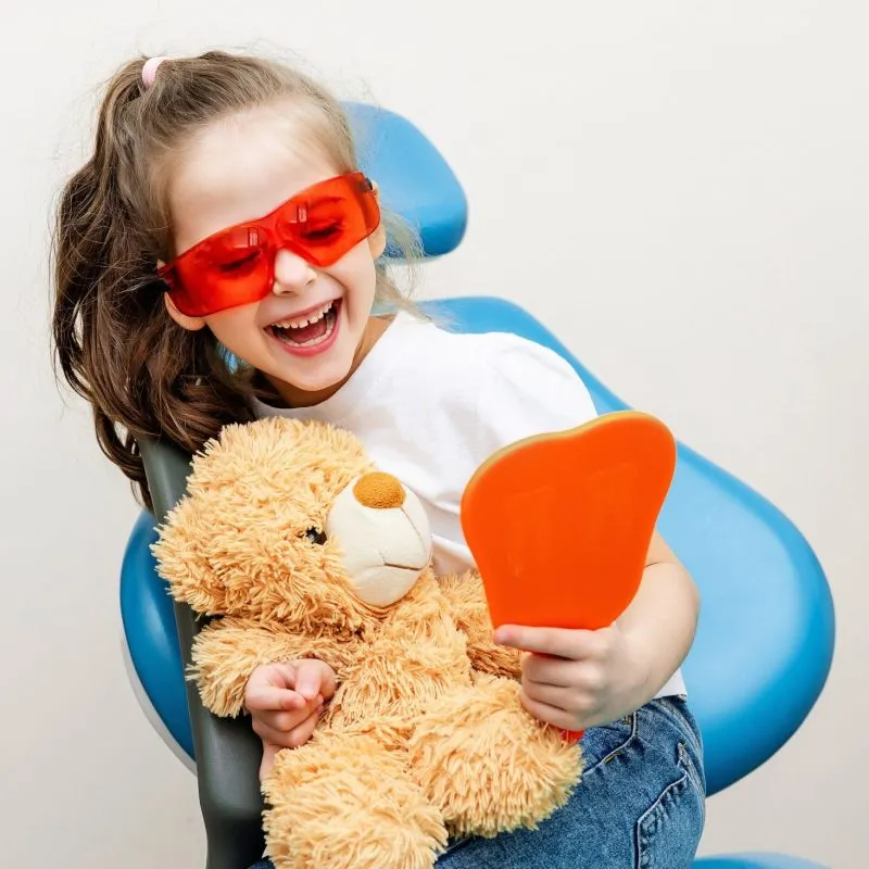 Young child smiling looking at her teeth in a mirror while holding a teddy bear